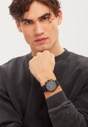 Ung mand med bølget brunt hår iført en mørk sweatshirt, hviler hagen på knyttet hånd og viser et sort armbåndsur med flere skiver og knapper.