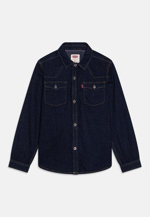 Mørkeblå denimskjorte med lange ærmer, to brystlommer med knapper, en krave og synlige gule syninger.