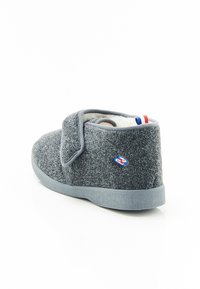 Grijze felt slipper met een klittenbandsluiting, witte pluche voering, laag ontwerp, rubberen zool en een klein blauw logo met rood/witte accenten.