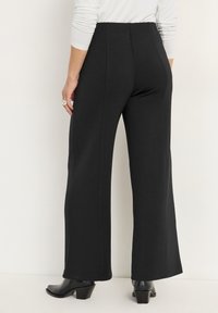 Zwarte wijd-uitlopende broek gemaakt van een gladde, rekbare stof. Het ontwerp heeft een hoge taille en rechte naden, gecombineerd met zwarte hakken.