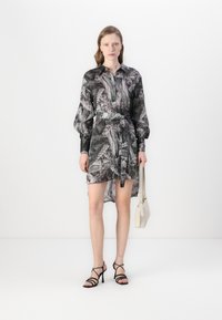 Abito camicia con una fantasia di foglie in nero e grigio, con maniche lunghe a sbuffo, vita cintrata da una cintura e orlo leggermente trasparente. Indossato con sandali neri con cinturini.