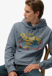 Sudadera de algodón gris con un diseño gráfico multicolor que presenta motivos florales y de dragones. Incluye un bolsillo frontal y cordones ajustables.
