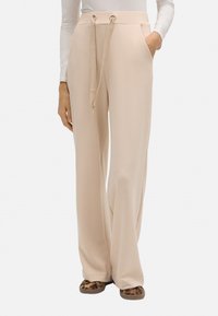 Pantalones de pierna ancha beige hechos de un tejido suave, con un cinturón de cordón, bolsillos laterales y una textura suave. Combinados con sandalias de estampado animal.