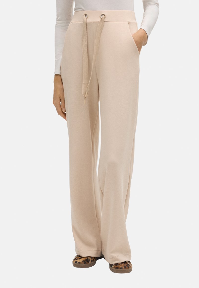 Pantalones de pierna ancha beige hechos de un tejido suave, con un cinturón de cordón, bolsillos laterales y una textura suave. Combinados con sandalias de estampado animal.