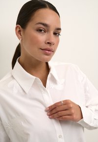 Camicia bianca con bottoni caratterizzata da ricami floreali sulle spalle. Modella che tocca delicatamente il colletto, mostrando un anello d'argento sulla mano.