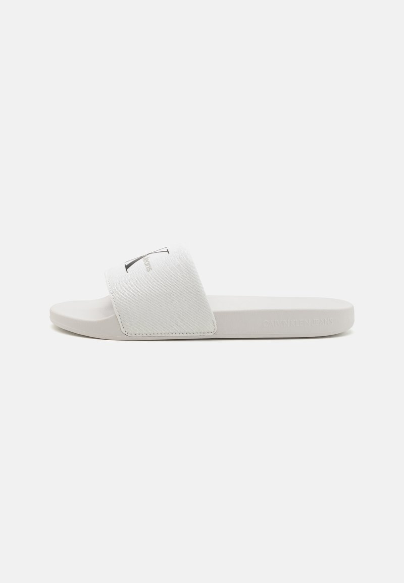 Hvite sklite sandaler med en stoffrem som har et logo. Sålen er glatt og flat, med et minimalistisk design og myk tekstur.