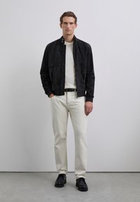 Mężczyzna stoi na jednolitym tle, ubrany w czarną zamszow�ą kurtkę, białą sweter, kremowe spodnie, czarny pasek i czarne buty, z rękami w kieszeniach.