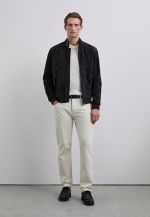 Man stands against plain background wearing black suede jacket, white sweater, cream pants, black belt, and black shoes, hands in pockets.