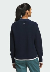Marineblauer Pullover mit geripptem Muster, versehen mit einem weißen Saum und Bündchen. Adidas-Logo ist am Nacken auf der Rückseite eingestickt.