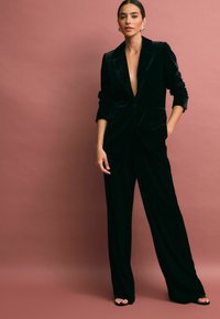 Svart sammetkostym med en djup V-ringad blazer i figurnära design och vida byxor, uppvisad mot en mjukt rosa bakgrund.