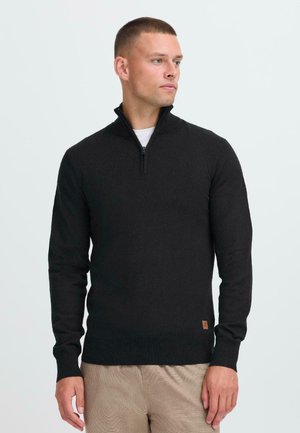 Black half-zip pullover sweater in a soft knit fabric, featuring long sleeves and a small logo patch at the hem. Worn over a white shirt.