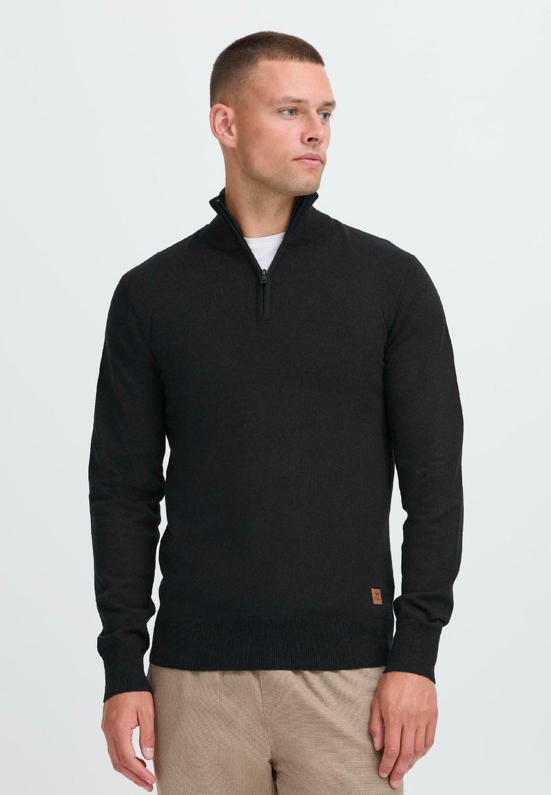 Pull-over noir à demi-zippé en tissu tricoté doux, avec des manches longues et un petit logo brodé à l'ourlet. Porté par-dessus une chemise blanche.