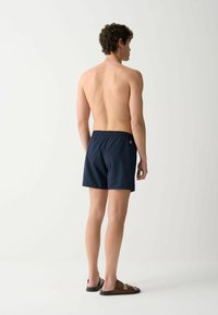 Costumi da bagno blu navy con vita elastica, tasca laterale e piccolo dettaglio con logo, abbinati a sandali aperti marroni, indossati da un modello maschile senza maglietta.