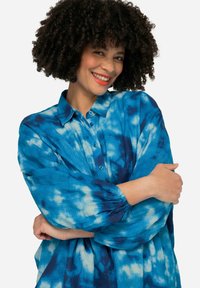 Blusa blu tie-dye con chiusura a bottoni, colletto a punta e maniche lunghe con polsini elasticizzati. Tessuto morbido e testurizzato con motivi simili a nuvole.