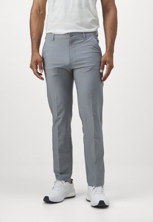 Άνδρας που φοράει ανοιχτό γκρι παντελόνι στενής γραμμής και λευκά αθλητικά παπούτσια, στέκεται μπροστά σε ένα απλό λευκό φόντο.