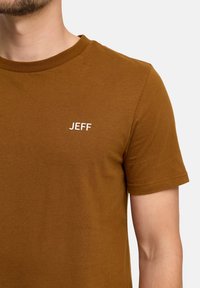 Brązowy t-shirt z krótkim rękawem z okrągłym dekoltem i białym logo "JEFF" na piersi, wykonany z miękkiej bawełny o gładkiej fakturze.