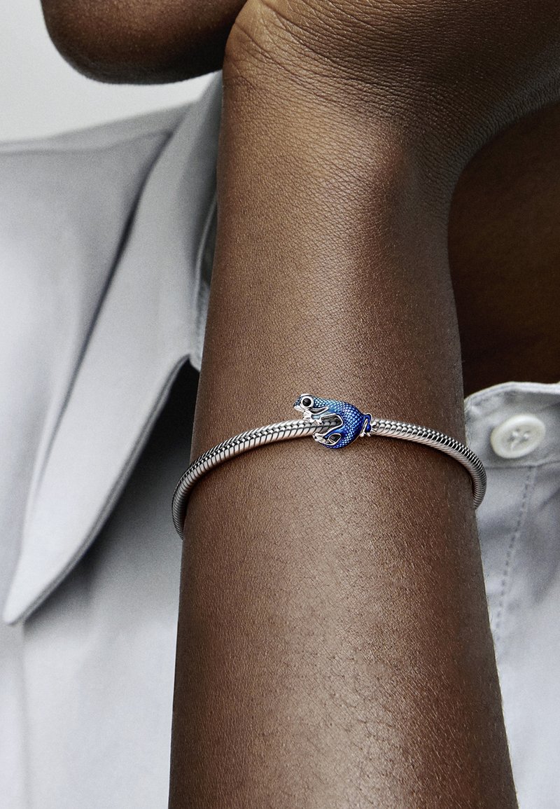 Strieborný náramok s rybím vzorom v modrých akcentoch. Textúrované a hladké povrchy, nosený na tmavej pleti na pozadí svetlej košele.