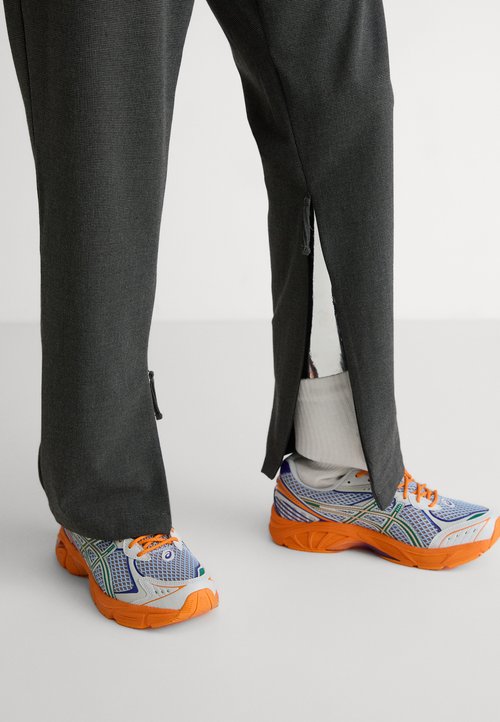 Pantalones atléticos grises con cremalleras laterales, combinados con zapatillas de correr multicolores que cuentan con una parte superior de malla y suelas de goma naranja.