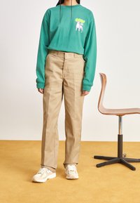 Grön långärmad skjorta med grafisk tryck, beige byxor och krämfärgade sneakers. I bakgrunden finns en gul matta och en trästol.