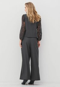 Woman standing facing away, wearing black lace-sleeve top, wide-leg black pants, and black high-heeled ankle boots.