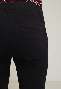 Czarne spodnie o dopasowanym kroju, z dwoma tylnymi kieszeniami i widocznymi detalami szwów. Tkanina wydaje się gładka z lekką teksturą.