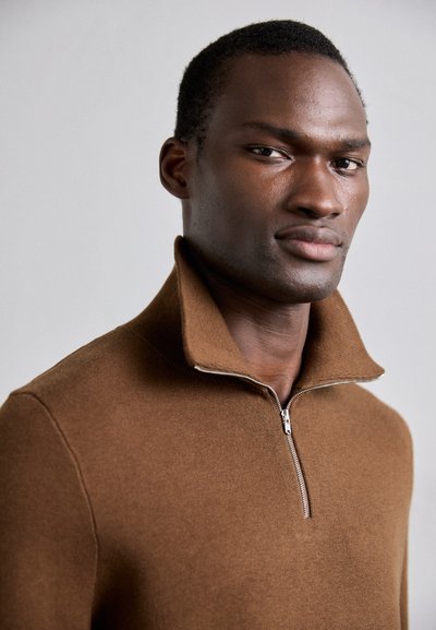 Pull en maille marron avec un col haut et des détails en zip, présentant une texture lisse et un design épuré. L'arrière-plan est sobre.