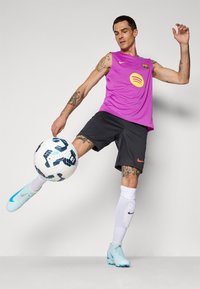Tetovált férfi lila FC Barcelona ujjatlan mezben és fekete rövidnadrágban, amint egy fehér-fekete focilabdát rúg benti környezetben.