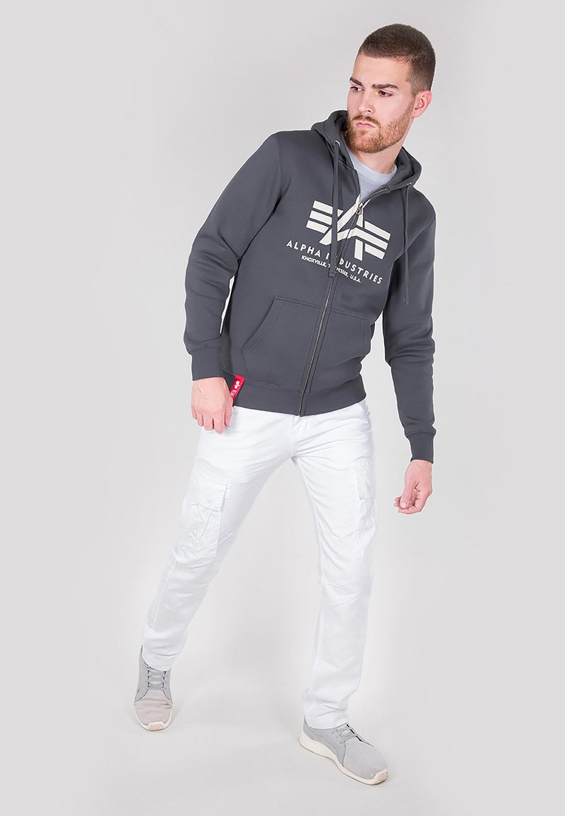 Šedá mikina na zips s logom, biele cargo nohavice a šedé tenisky. Hladká látka a strih s prednými vreckami a kapucňou na šnúrku.