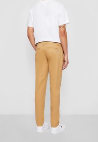 Beige chinos med slim fit, med bakfickor och en logodetalj nära midjan, tillsammans med en enkel vit t-shirt och vita skor.