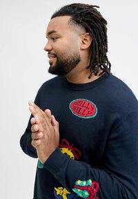 Navy-blue sweater featuring knitted holiday-themed designs, including "Holly Jolly" text, gifts, and festive accents in bright colors.