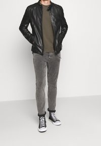 Schwarze Lederjacke, olivgrünes T-Shirt, graue Slim-Fit-Jeans und schwarze High-Top-Sneaker mit weißen Sohlen. Einfaches Design und figurbetonte Passform.