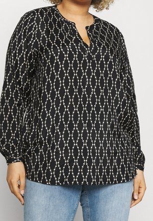 Sort damebluse med lange ærmer og hvidt prikket diamantmønster, delt udskæring, båret med lysblå jeans.