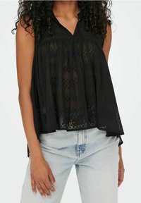 Mustat hihattomat paita, joka on valmistettu kevyestä, läpinäkyvästä kankaasta, jossa on kohokuvioitu kukkakuosi ja kerrostettu muotoilu, yhdistettynä vaaleansinisiin farkkuihin.