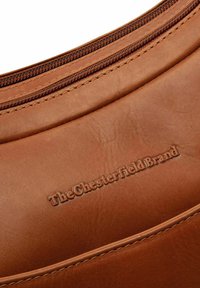Bruine leren tas met een gladde textuur, gebogen ontwerp, zichtbare stiksels en een geperste logo met de tekst "The Chesterfield Brand" bij de rits.