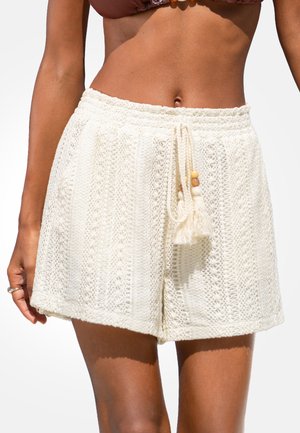 Nahaufnahme einer Person, die weiße, gehäkelte Shorts mit hohem Bund, Kordelzug und Quasten trägt, kombiniert mit einem braunen Oberteil.