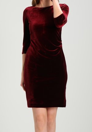 Femme portant une robe en velours rouge foncé, ajustée, longueur genou, avec manches trois-quarts, sur un fond clair uni.