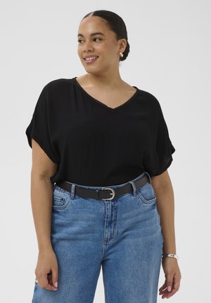 Femme souriante, portant un chemisier noir à manches courtes en V rentré dans un jean bleu taille haute avec une ceinture noire à boucle argentée.