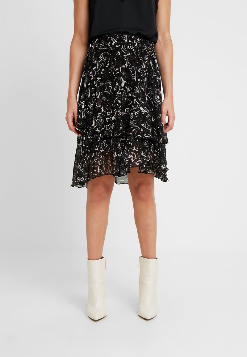 Femme portant une jupe noire à fleurs en plusieurs couches avec un haut noir et des bottines blanches, debout devant un fond clair uni.