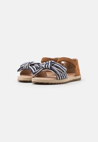 Bruna mockaskinnsandaler med en marinblå och vit randig textilrosett. Har en vävd sula i jute och justerbart rem för en säker passform.