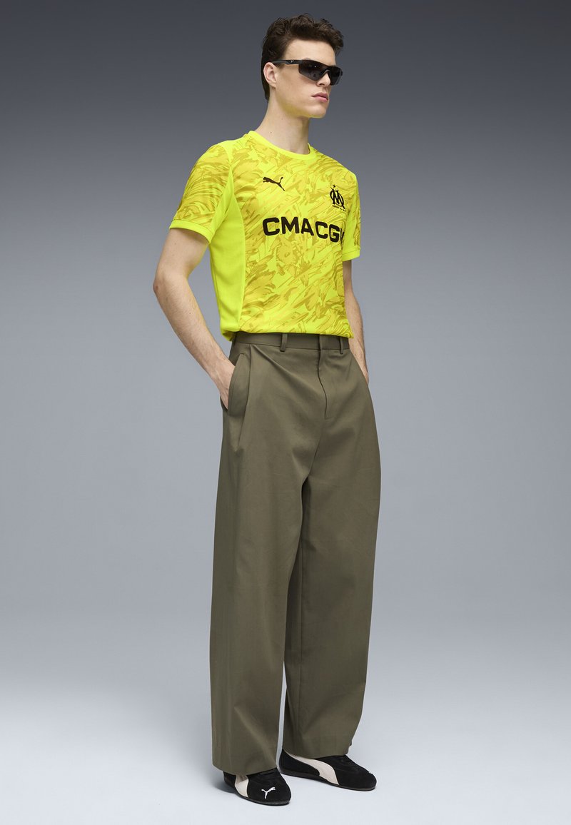 Jasnożółta koszulka sportowa z wzorem kamuflażu, czarnym napisem, połączona z luźnymi oliwkowymi spodniami i czarnym obuwiem.