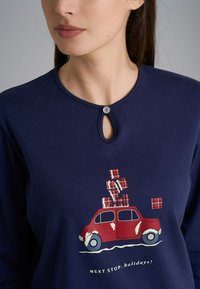 Maglietta a maniche lunghe blu navy con scollatura a chiave, decorata con un grafiche di una macchina rossa con regali impilati in un motivo a quadretti, e la scritta "PROSSIMA FERMATA: festività!" sotto.