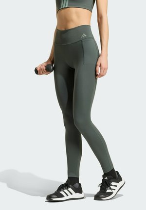 Frau trägt grüne Leggings mit hoher Taille und Sport-BH, hält eine schwarze Hantel, steht in schwarz-weißen Adidas-Sneakers vor hellem Hintergrund.