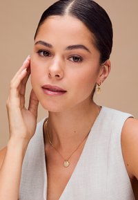Collana con pendente a forma di conchiglia in oro su una delicata catena, indossata con piccoli orecchini a cerchio in oro, abbinata a un capo leggero e strutturato.