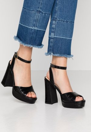 Sandales plateformes noires avec une texture embossée effet croco, dotées d'un talon épais et d'une bride cheville, associées à un pantalon en denim bleu coupé avec ourlets effilochés.