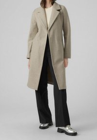 Lichtbeige jas met een notch revers, rechte snit en lange mouwen. Draagtt over een geribbeld shirt, gecombineerd met zwarte broek en glanzende zilveren schoenen.