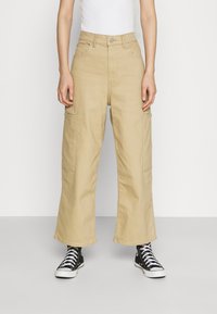 Béžové cargo kalhoty s širokým střihem, které mají dvě boční kapsy a zapínání na knoflík, nošeny s černými vysokými teniskami.