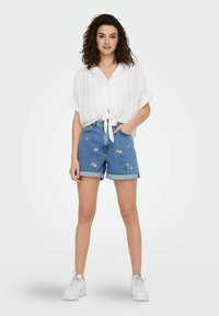 Chemise blanche à boutons avec un devant noué, assortie à un short en denim bleu avec des broderies de marguerites. Le mannequin porte des baskets blanches.