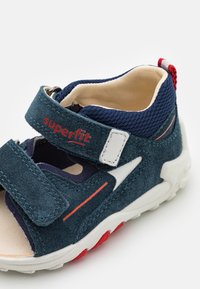Blue suede sandal with mesh accents, two Velcro straps, and red detailing. White sole featuring textured grip. Lightweight and flexible design.