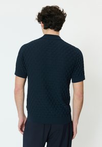 Moški s kodrastimi lasmi, oblečen v temno modro polo majico s kratkimi rokavi iz teksturirane pletenine, viden od zadaj na enostavni svetli podlagi.