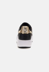 Black sneaker with gold accent at the heel, featuring a textured upper and a white rubber sole. Logo displayed on the back.